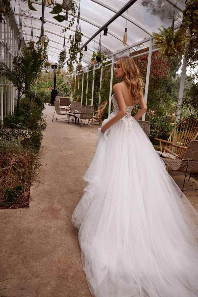 Eine Frau in einem trägerlosen, fließenden weißen Brautkleid geht einen mit Pflanzen und leeren Stühlen gesäumten Gewächshausgang hinunter, während das Sonnenlicht durch die Glasdecke fällt.