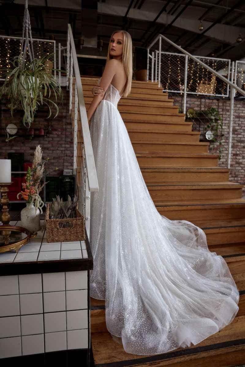 Eine Frau in einem funkelnden, trägerlosen weißen Brautkleid mit langer Schleppe steht auf Holztreppen, blickt elegant über die Schulter in einem modernen Indoor-Setting mit Pflanzen und Lichterketten - ein stilvolles Beispiel für aktuelle Brautmoden.