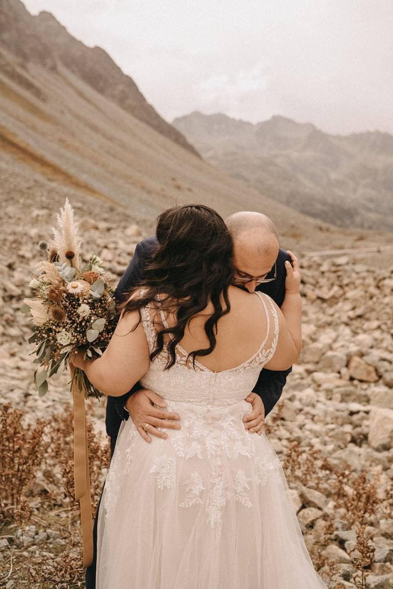 Ein Paar umarmt sich im Freien auf felsigem Terrain, Berge im Hintergrund. Die Person in einem weißen Brautkleid hält einen Blumenstrauß, während ihr Partner im dunklen Anzug sie innig umarmt. Perfekt für Brautmoden-Inspiration.