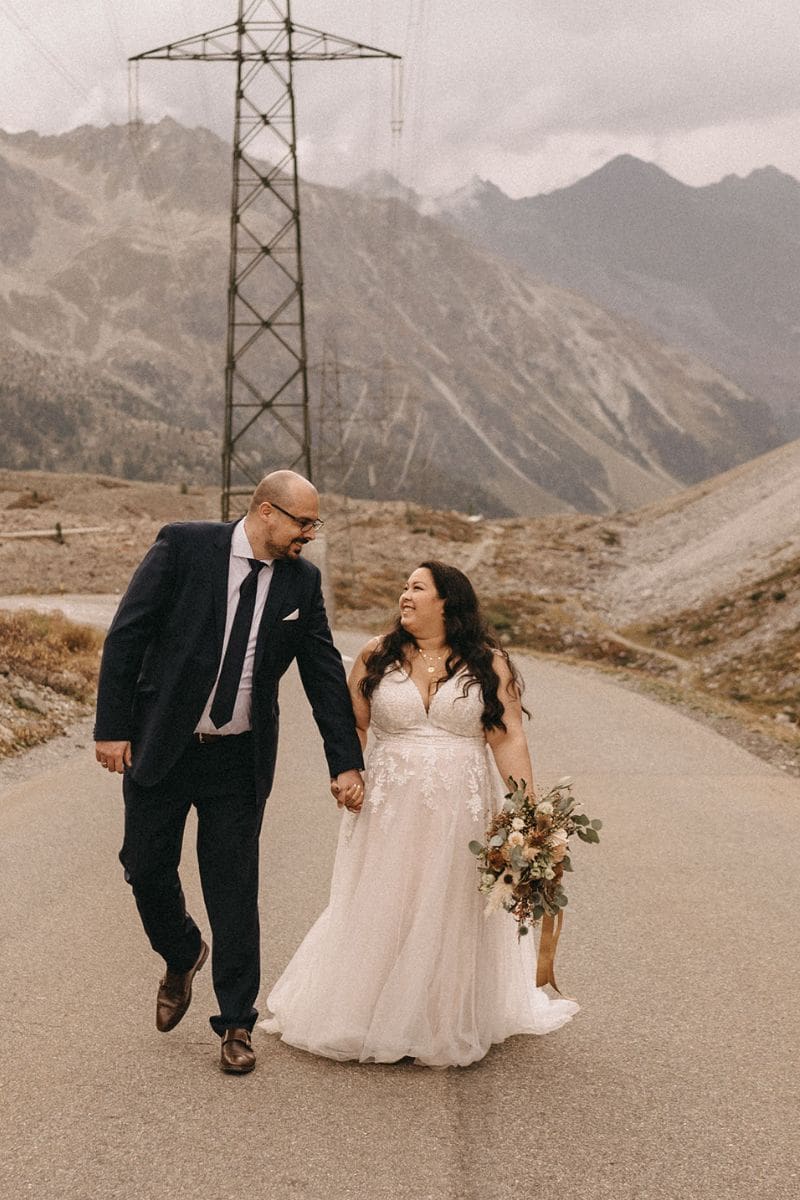 Ein frisch verheiratetes Paar spaziert Hand in Hand auf einer Bergstraße und lächelt sich gegenseitig an. Der Bräutigam trägt einen dunklen Anzug, und die Braut strahlt in einem Brautkleid und hält ihren Blumenstrauß in der Hand. Im Hintergrund sind felsige Berge und Stromleitungen zu sehen.