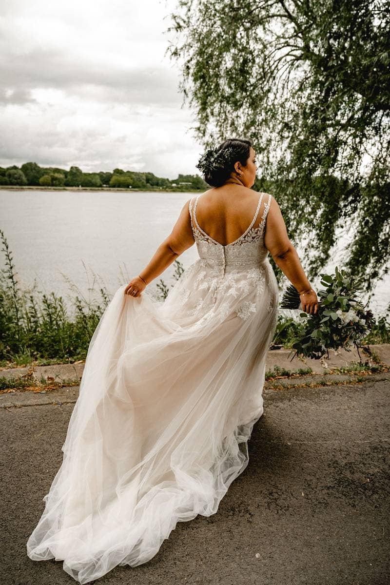 Eine Braut in einem wallenden Hochzeitskleid hält mit der einen Hand ihr Kleid und mit der anderen einen Blumenstrauß hoch und schreitet einen von Grün und Bäumen gesäumten Uferweg entlang.
