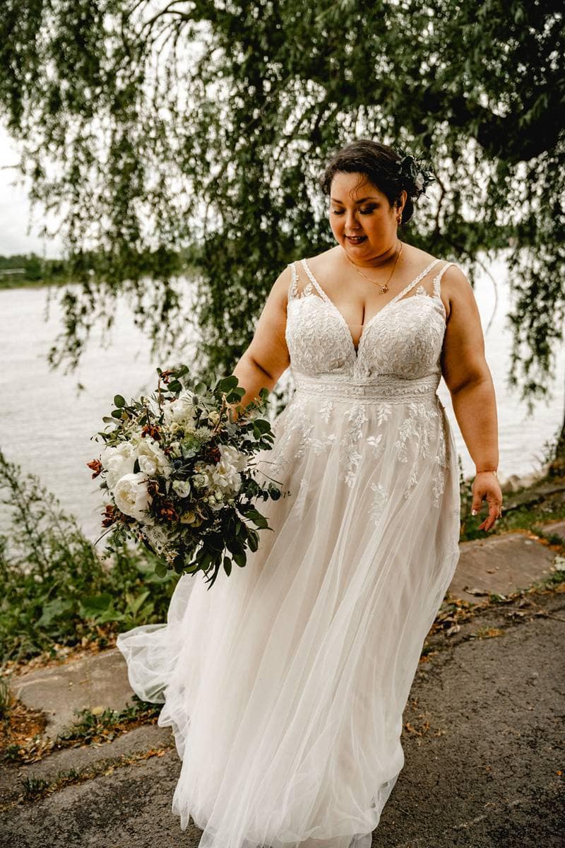 Eine Braut in einem spitzenbesetzten Brautkleid mit einem Strauß weißer und grüner Blumen geht einen Uferweg entlang, mit Bäumen und Wasser im Hintergrund.