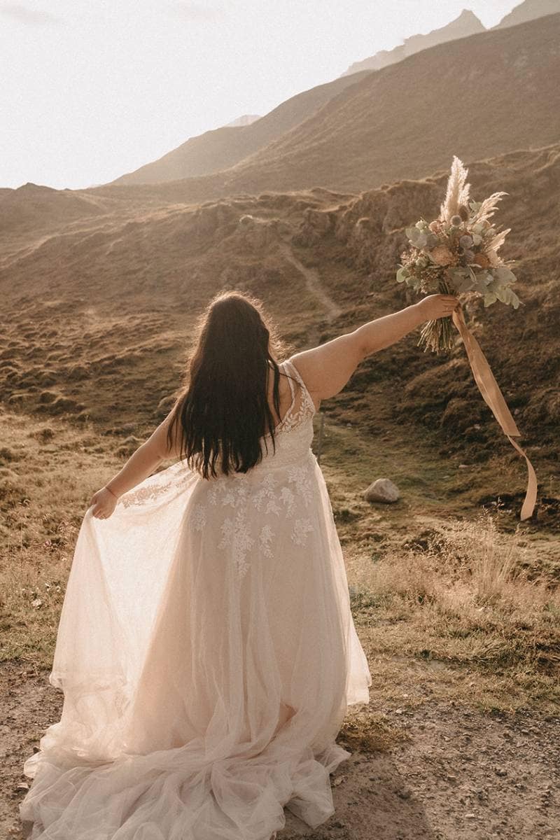 Eine Braut in einem zarten Brautkleid steht im Freien auf einem sonnenbeschienenen Hügel, hält ihren Brautstrauß hoch und hebt ihr Kleid, während sie von der Kamera weg in Richtung der Berge blickt.
