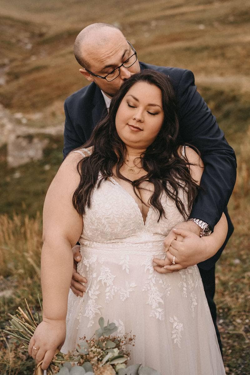 Ein Mann in einem dunklen Anzug umarmt liebevoll eine Frau in einem weißen Brautkleid von hinten. Sie stehen im Freien auf einer Wiese, beide wirken ruhig und zufrieden, während die Braut einen kleinen Blumenstrauß hält.