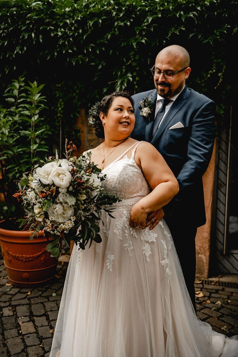 Eine Braut in einem atemberaubenden Hochzeitskleid hält einen großen Blumenstrauß, während sie mit ihrem Bräutigam in einem dunklen Anzug steht. Sie lächeln und umarmen sich im Freien, umgeben von üppigem Grün und kopfsteingepflasterten Wegen.