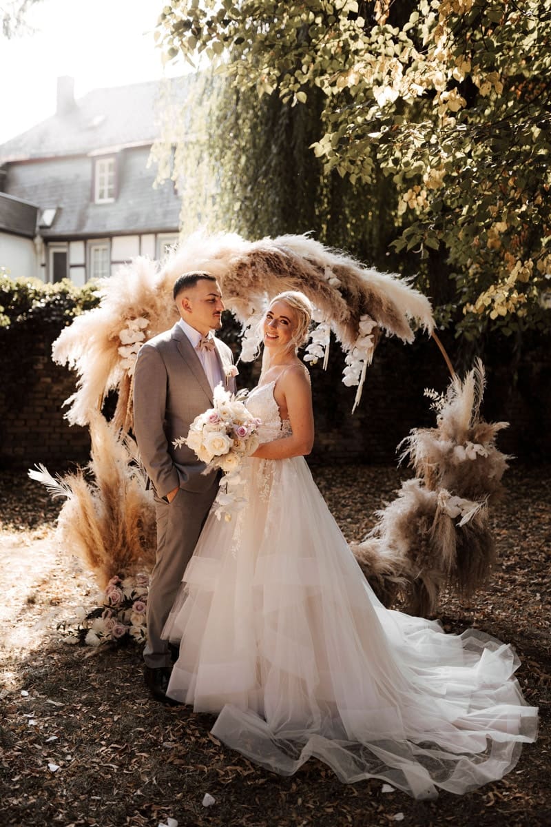 Eine Braut in einem fließenden weißen Brautkleid und ein Bräutigam in einem hellgrauen Anzug stehen lächelnd unter einem Bogen aus Pampasgras im Freien, umgeben von Grünzeug und gedämpftem Sonnenlicht - ein bezaubernder Moment von Brautmoden-Eleganz.