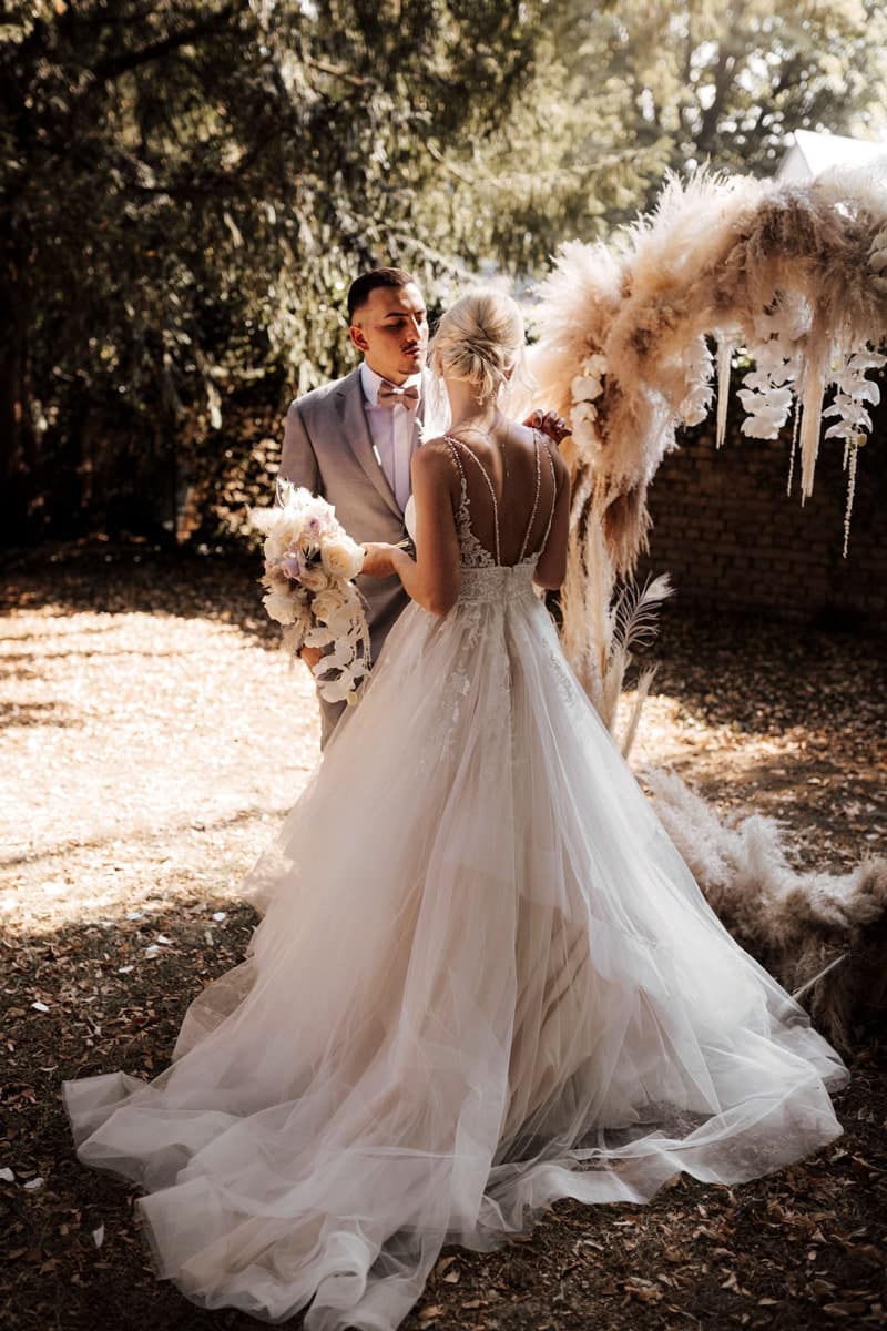Eine Braut in einem fließenden weißen Brautkleid und ein Bräutigam in einem hellen Anzug stehen dicht beieinander im Freien unter einem Blumenbogen, umgeben von Bäumen und Sonnenlicht, das durch die Blätter fällt.