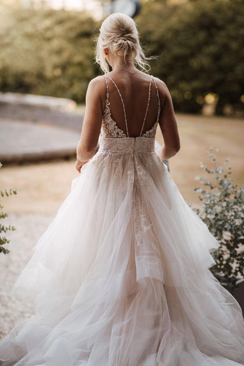 Eine Frau mit blondem Haar in einer Hochsteckfrisur trägt ein umwerfendes weißes Hochzeitskleid aus Spitze und Tüll mit dünnen Trägern, steht draußen vor viel Grün und einem Steinweg. Ihr elegantes Brautkleid fängt den Zauber moderner Brautmoden ein.