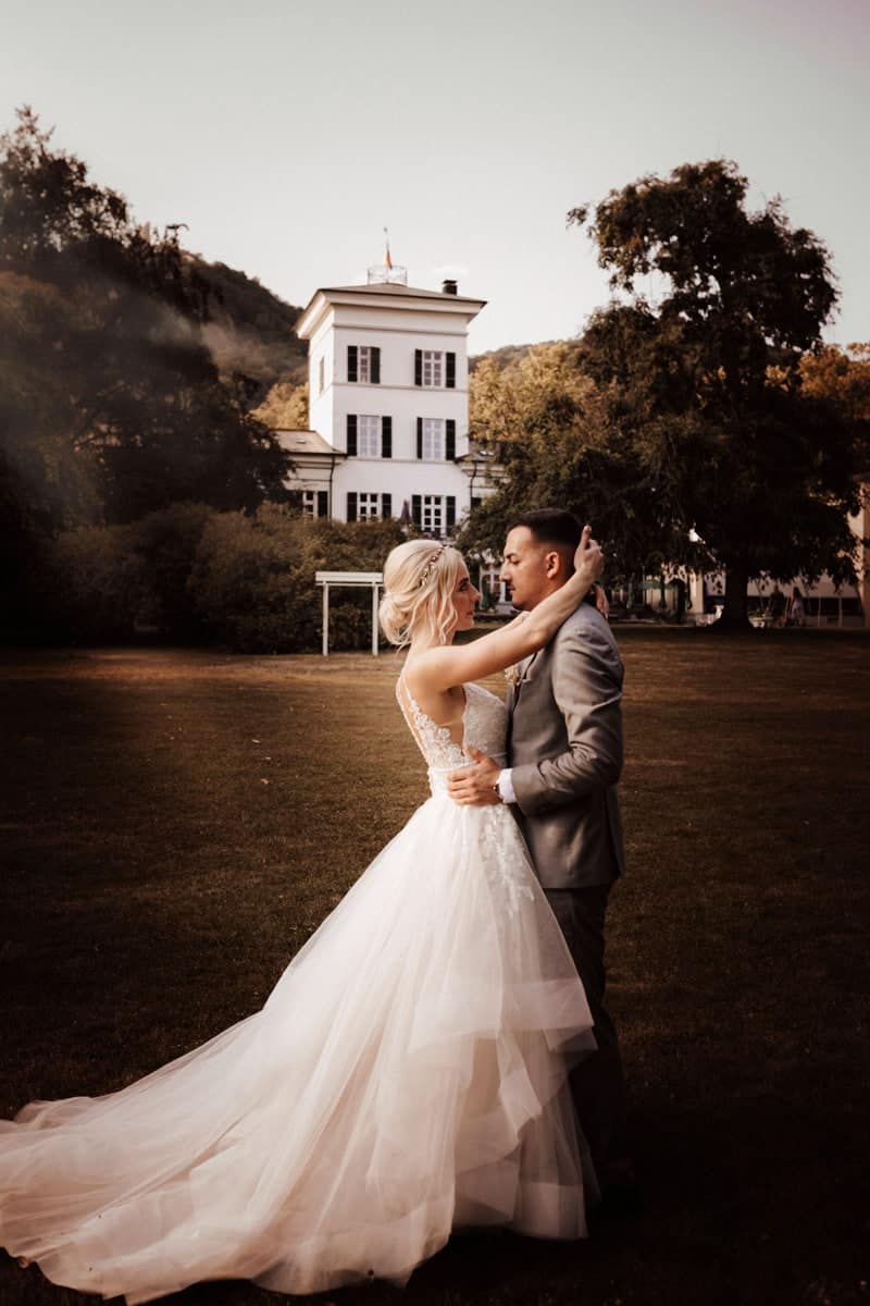 Eine Braut in einem atemberaubenden Hochzeitskleid und ein Bräutigam in einem grauen Anzug umarmen sich auf einem Rasen vor einem großen weißen Gebäude und Bäumen. Diese Hochzeitsszene im Freien bei Sonnenuntergang fängt pure Romantik ein.