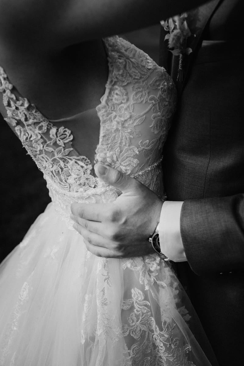 Ein Schwarz-Weiß-Foto in Nahaufnahme von einer Umarmung zwischen Braut und Bräutigam. Die Hand des Bräutigams ruht sanft auf dem Brautkleid und hebt die zarten Spitzendetails des Hochzeitskleids und seines Anzugs hervor. Die Gesichter der beiden sind nicht zu sehen.