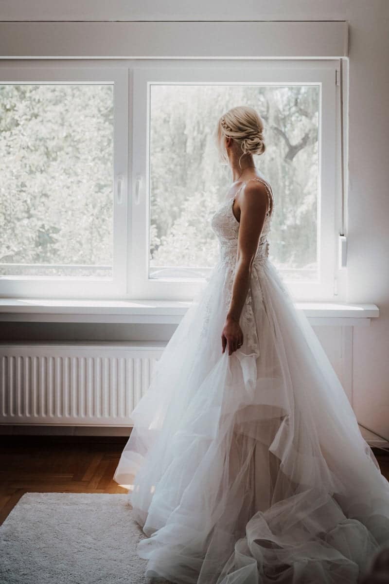 Eine Braut in einem weißen Brautkleid steht drinnen an einem großen Fenster und blickt nach draußen. Sanftes natürliches Licht beleuchtet ihr Hochzeitskleid, und ihr blondes Haar ist zu einer eleganten Hochsteckfrisur gestylt.