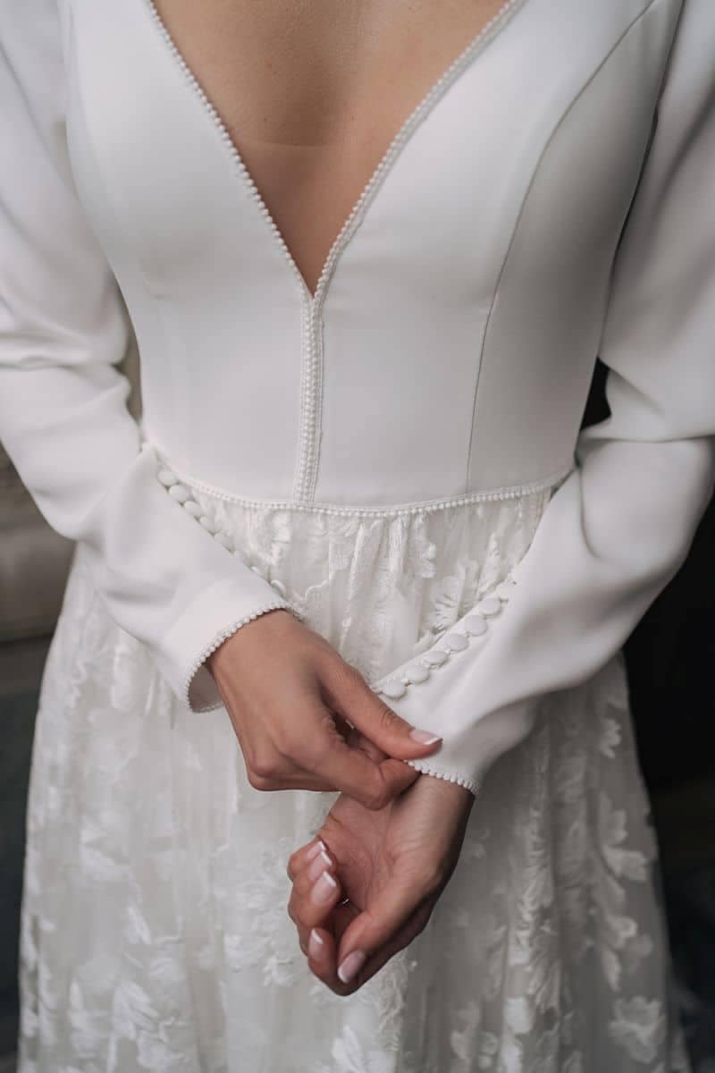 Dama 2021 Eine Frau in dem Dama Couture Latifa Hochzeitskleid mit langen Ärmeln und tiefem V-Ausschnitt rückt die geknöpfte Manschette am Ärmel zurecht. Das taillierte Mieder und der Rock aus floraler Spitze unterstreichen den raffinierten Stil dieses eleganten Brautkleides.