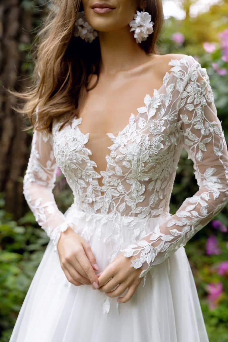 Eine Frau, die das Dama Couture Calista Brautkleid trägt - ein langärmeliges, florales Spitzenhochzeitskleid mit tiefem V-Ausschnitt - steht in einem Garten, umgeben von üppigem Grün und rosa Blumen.