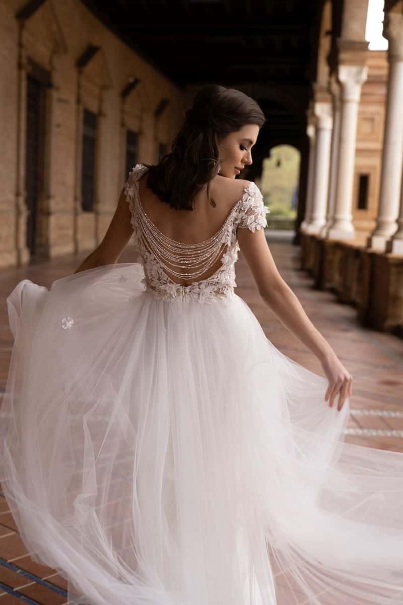Dama Couture Seville Eine Frau im Dama Couture Pavetta Brautkleid mit fließender weißer Silhouette, tiefem Rücken und Perlenketten steht auf einem überdachten Spaziergang im Freien, blickt über ihre Schulter und hält ihren Rock fest - moderne Braut-Eleganz.