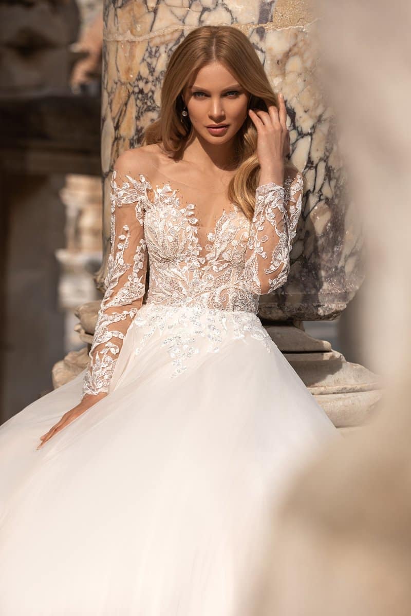 Oliver Martino Rosanna Brautkleid Eine Frau posiert neben einer Marmorsäule, berührt ihr Haar und schaut in die Kamera. Sie trägt das Oliver Martino Rosanna Brautkleid, ein langärmeliges Hochzeitskleid aus Spitze und Tüll mit Blumendetails.