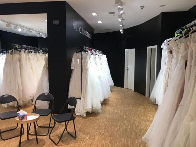 Ein Brautmodengeschäft mit weißen Brautkleidern, die an den Wänden hängen, zwei schwarzen Klappstühlen und einem kleinen runden Tisch mit Tüchern in der Mitte eines Holzbodens.