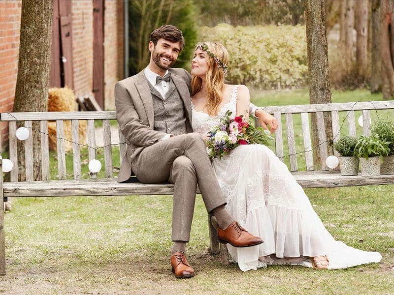 Eine Braut in weißem Kleid und Blumenkranz sitzt auf einer Holzbank, hält einen Blumenstrauß und lächelt den Bräutigam in einem hellbraunen Anzug von hochzeitsrausch Men an. Sie stehen im Freien zwischen Bäumen und einem Gebäude und sehen glücklich und entspannt aus.