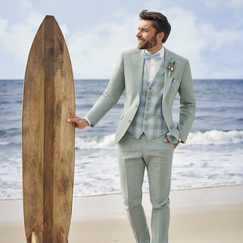 Ein Mann in einem hellgrünen Anzug steht mit einem hölzernen Surfbrett am Strand und lächelt. Er wartet auf seine Braut, die beide mit der Atelier Torino Vintage Wedding Schleife / Tuch 821779/30 feiern.