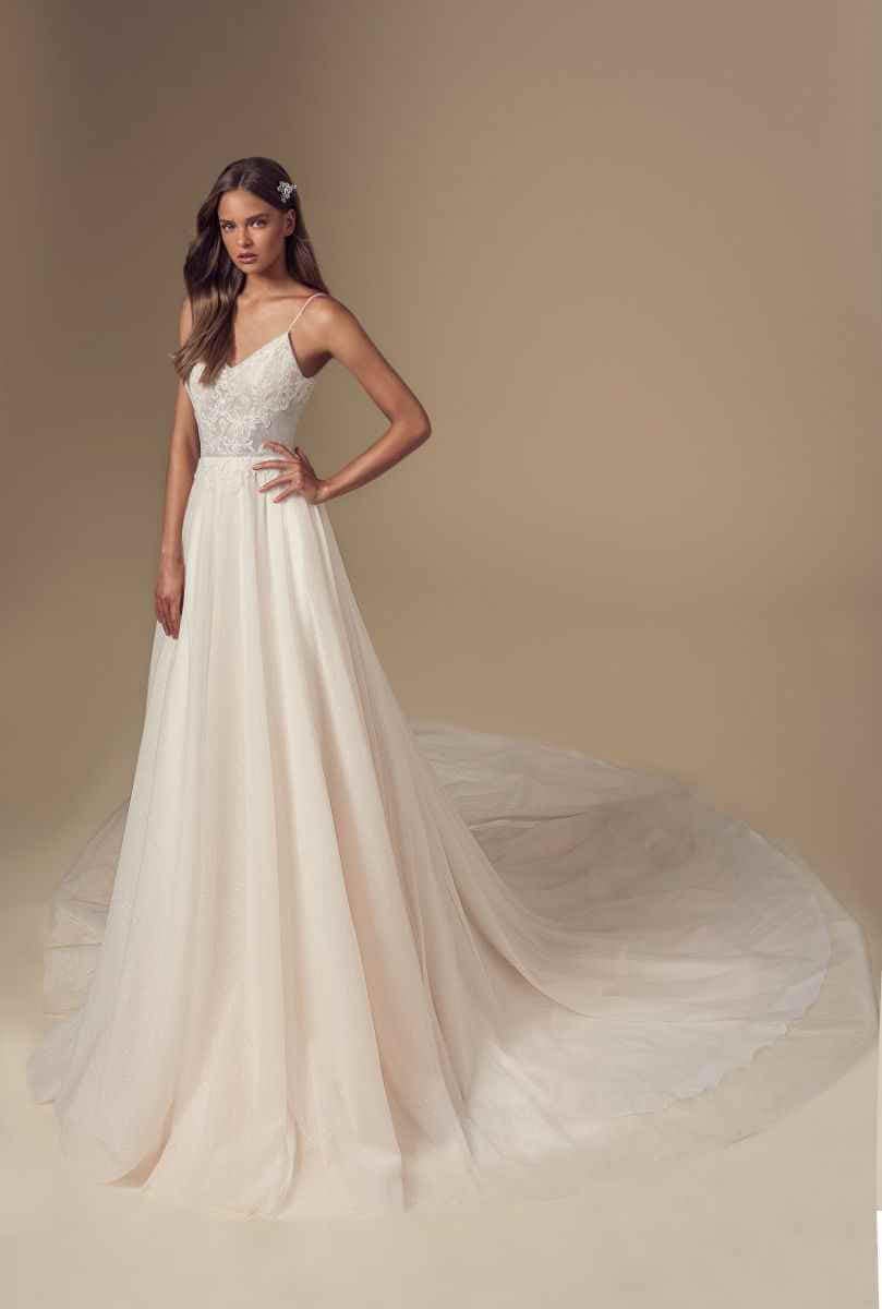 Eine Frau steht in einem Atelier und trägt ein langes, fließendes weißes Brautkleid mit dünnen Trägern, Spitzenmieder und einer langen Schleppe. Sie hat langes, gewelltes Haar und ein Haar-Accessoire und posiert anmutig vor einem beigen Hintergrund.