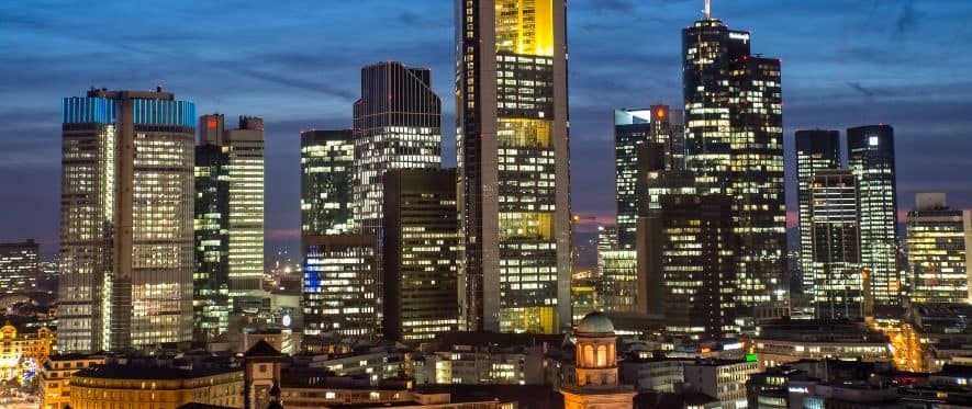 Eine Stadtsilhouette in der Abenddämmerung mit hohen, beleuchteten Wolkenkratzern und Büroleuchten kontrastiert mit einem historischen Gebäude im Vordergrund, in dem eine Braut in einem eleganten Brautkleid vor dem tiefblauen Himmel posiert.