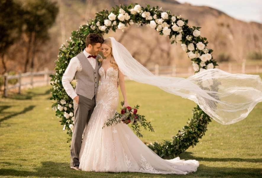 Eine Braut in einem atemberaubenden Brautkleid und ihr Bräutigam stehen im Freien vor einem runden Blumenbogen. Ihr Schleier weht im Wind, während sie lächeln, umgeben von üppigem Grün und einem klaren Himmel - ein bezaubernder Moment der Liebe und Brautmoden-Eleganz.