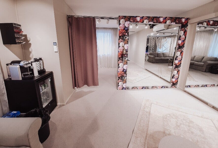 Ein geräumiger, sanft beleuchteter Raum mit beigem Teppich, floral gerahmten und schlichten Spiegeln an einer Wand, einem vorgehangenen Bereich zum Anprobieren von Brautkleid oder Hochzeitskleid, einem Sofa und einer Kaffeestation mit einer Kaffeemaschine und Zubehör.