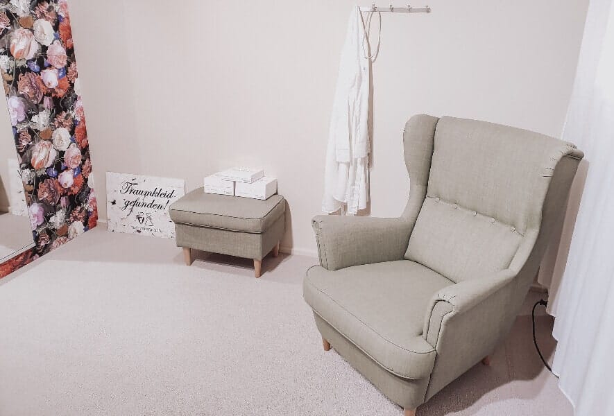 Ein gemütlicher Raum mit einem hellgrünen Sessel und einem Ottomanen, einem geblümten Vorhang und einem weißen Bademantel - perfekt, um Brautmoden anzuprobieren oder sich zu entspannen, bevor man in sein Hochzeitskleid schlüpft. An der hellen Wand stehen Schachteln und ein dekoratives Schild.