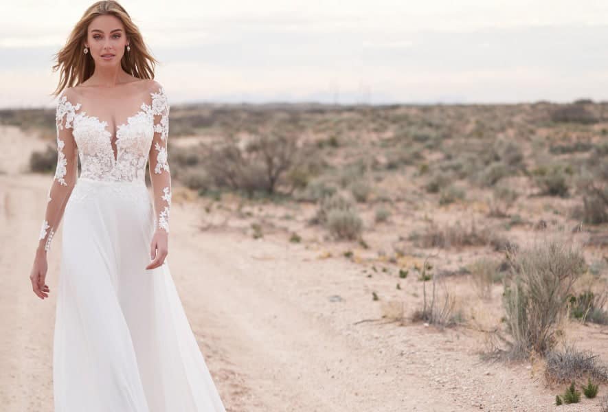 Eine Frau in einem langärmeligen weißen Hochzeitskleid geht einen unbefestigten Weg in einer Wüstenlandschaft entlang, mit spärlicher Vegetation und einem bewölkten Himmel im Hintergrund.