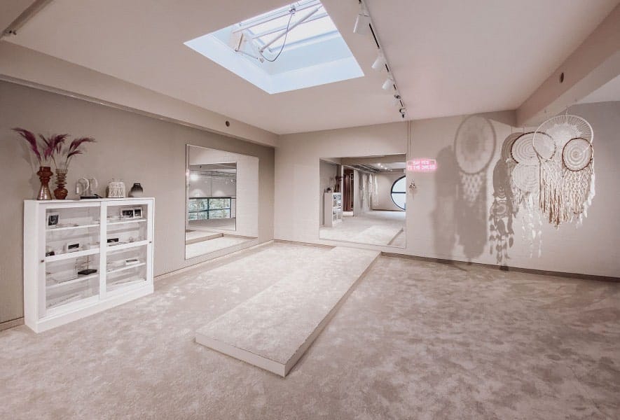 Ein geräumiger, sanft beleuchteter Raum mit hellem Teppich, einem Oberlicht, Wandspiegeln, weißen Regalen und großen Traumfängern. Minimales Dekor und eine beruhigende, neutrale Farbpalette machen ihn zu einem idealen Ort, um Ihr perfektes Brautkleid auszusuchen oder nach Brautmoden zu stöbern.