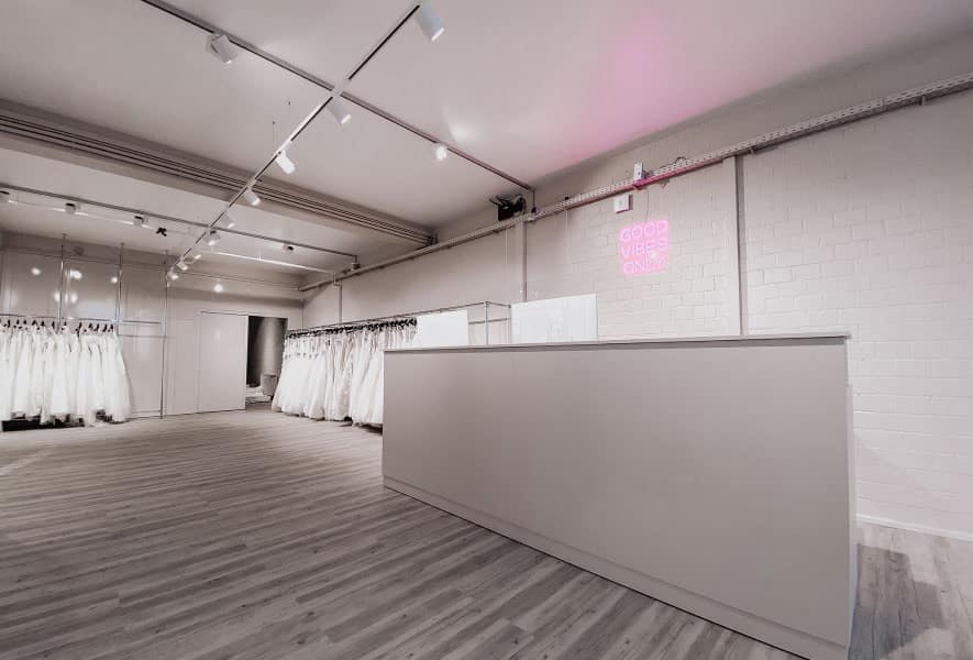 Ein minimalistisches Brautmodengeschäft mit eleganten Brautkleidern und Brautmoden in der Auslage, einem modernen weißen Empfangstresen, hellem Holzfußboden und einem rosa Neonschild an der Wand mit der Aufschrift GOOD VIBES ONLY.