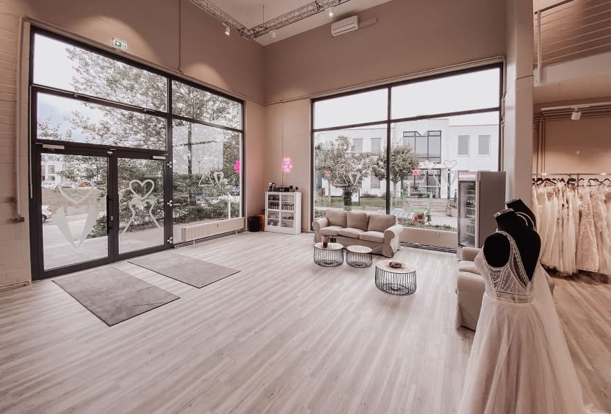 Eine geräumige, moderne Brautmoden-Boutique mit großen Fenstern, mehreren exquisiten Brautkleid-Varianten in der Auslage, einer gemütlichen Sitzecke mit Sofas und Couchtischen und hellem Holzfußboden. Natürliches Licht durchflutet den Raum.