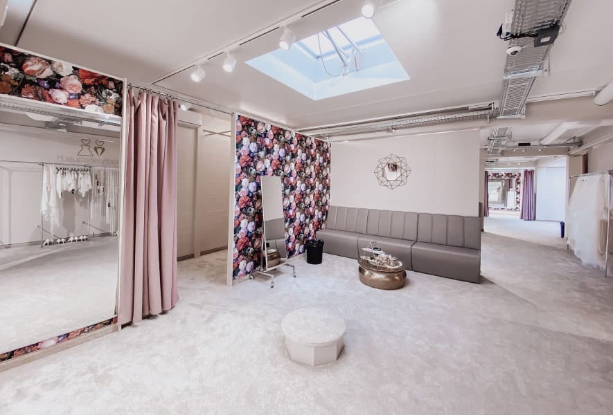 Eine moderne, elegante Umkleidekabine mit Blumentapete, einem grauen Sofa, rosa Vorhängen, einem runden Couchtisch, Spiegel und hellem Oberlicht. Weißes Brautkleid hängt im Hintergrund an einem Kleiderständer.