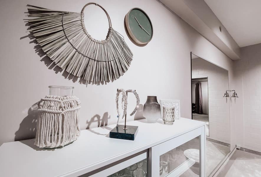 Ein moderner weißer Konsolentisch mit dekorativen Elementen, darunter ein Makramee-Gefäß, eine abstrakte Skulptur, eine strukturierte Vase und Glaswaren. Darüber befinden sich eine runde Wanduhr und fächerförmige Wandkunst. Ein großer Spiegel reflektiert die minimalistische Brautkleid-Ausstellung.