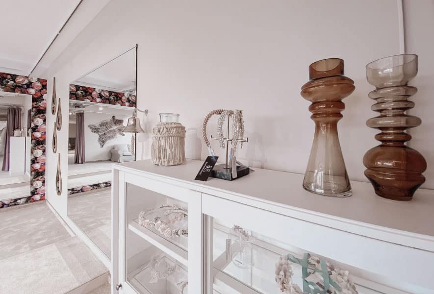 Auf einer weißen Anrichte stehen Glasvasen, Schmuck und Dekorationsartikel. Ein großer Spiegel und eine Blumentapete akzentuieren den modernen, sanft beleuchteten Raum - perfekt, um Brautmoden zu präsentieren oder das ideale Brautkleid auszuwählen.
