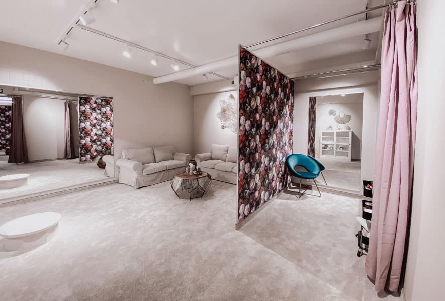 Ein geräumiger, moderner Anproberaum mit hellem Plüschteppich, geblümten Akzentwänden, einem beigen Sofa, einem blauen Stuhl, einem großen Spiegel und rosa Vorhängen - perfekt, um in gemütlicher und eleganter Atmosphäre Brautmoden anzuprobieren oder Ihr Traumhochzeitskleid zu finden.