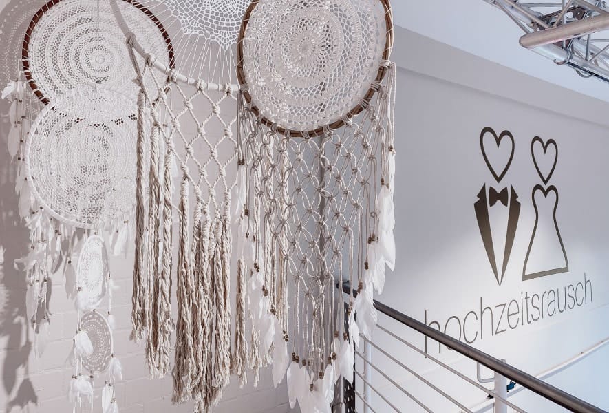 Große gehäkelte Traumfänger mit weißen Federn und Quasten hängen im Treppenhaus neben einer Wand mit einem stilisierten Brautpaar und dem Schriftzug "hochzeitsrausch" und stimmen auf die Wahl des Brautkleides oder des Brautmodens ein.