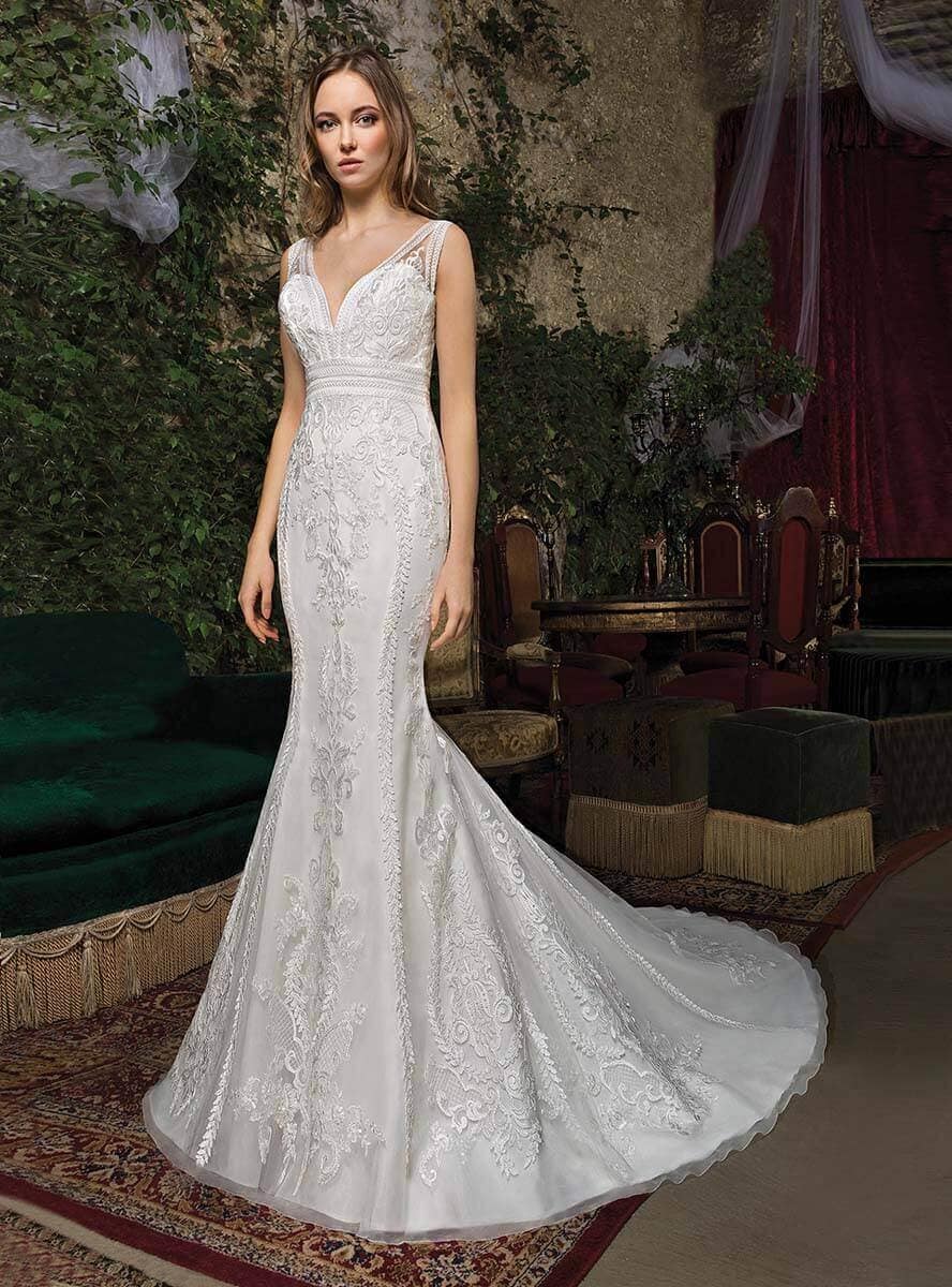 Eine Frau in einem taillierten, ärmellosen weißen Spitzen-Hochzeitskleid mit tiefem V-Ausschnitt steht in einem Innenraum auf einem gemusterten Teppich, umgeben von Samtmöbeln und verzierten Stühlen, ihr Look wird durch elegante Brautaccessoires perfekt ergänzt.