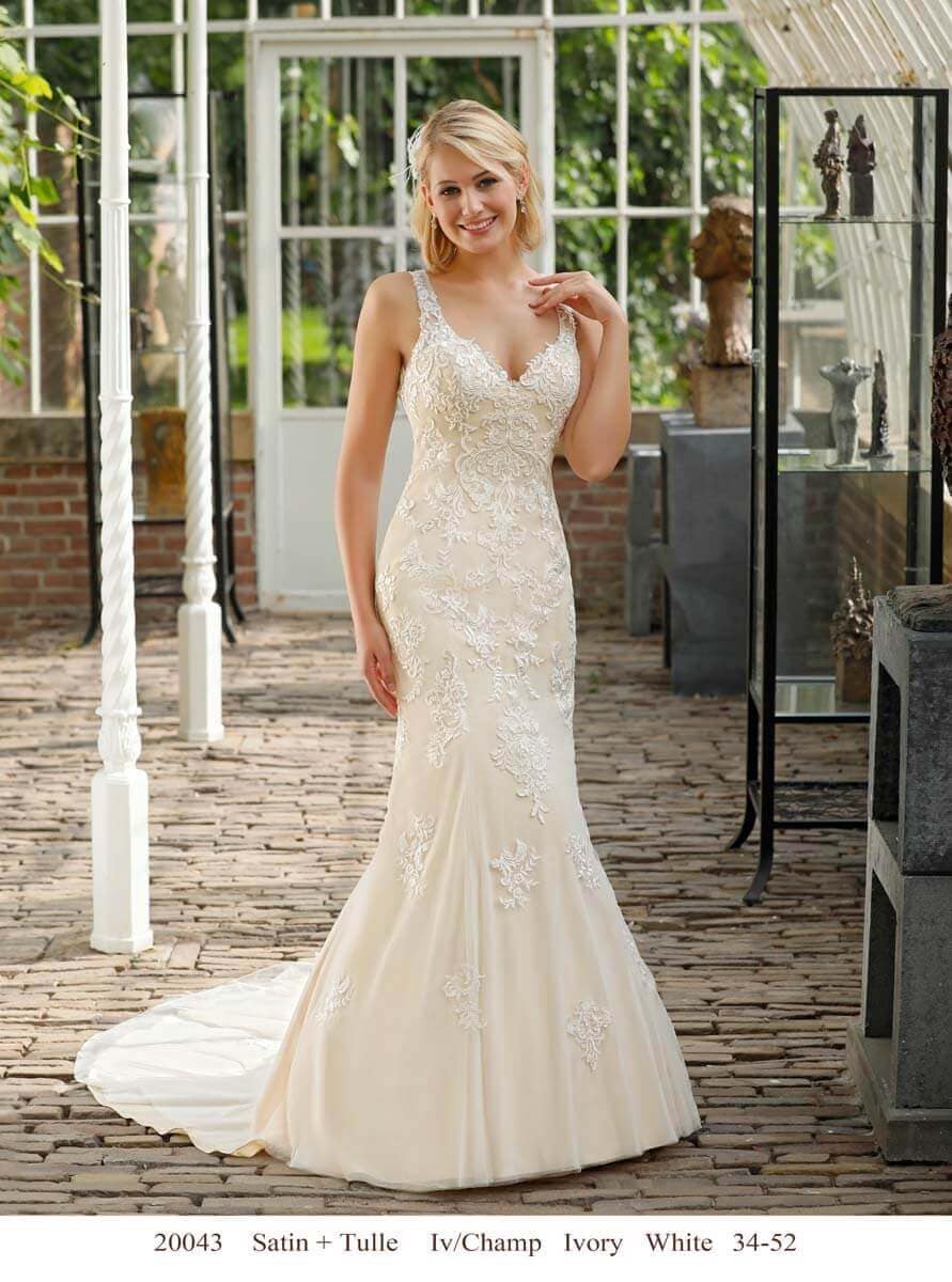 Eine Amelie-Braut in einem elfenbeinfarbenen Hochzeitskleid aus Satin und Tüll mit floralen Spitzendetails steht lächelnd in einem sonnendurchfluteten Gewächshaus mit Ziegelfußboden und Glaswänden.