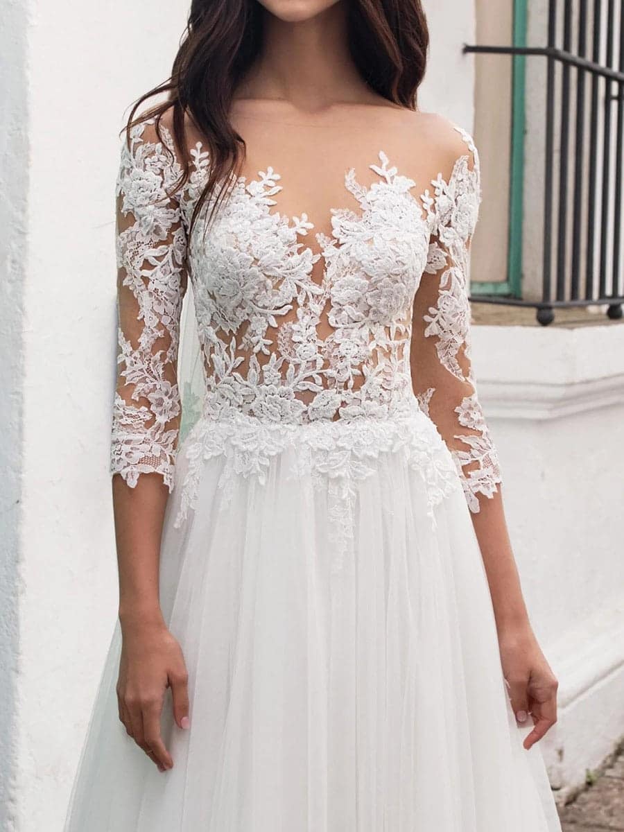 Pronovias-Fornax-2 Eine Frau in einem Fornax-Brautkleid von Pronovias mit durchsichtigen langen Ärmeln und aufwändiger Blumenspitze am Mieder und an den Ärmeln, die im Freien vor einer weißen Wand steht.