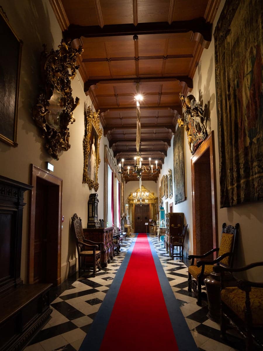 Ein prachtvoller Flur mit einer Holzdecke, kunstvollen Goldverzierungen, Kronleuchtern, antiken Möbeln und einem roten Teppich über einem schwarz-weiß karierten Boden - ideal, um Brautmoden oder ein elegantes Hochzeitskleid zu präsentieren. Wandteppiche und Gemälde säumen die Wände.