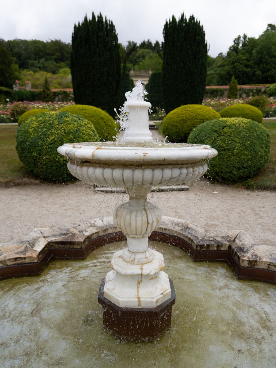 Ein weißer Marmorbrunnen versprüht Wasser in einem Landschaftsgarten mit gestutzten grünen Sträuchern, blühenden Blumen und hohen Hecken - eine ideale Kulisse für ein Brautkleid oder ein elegantes Hochzeitskleid-Fotoshooting.