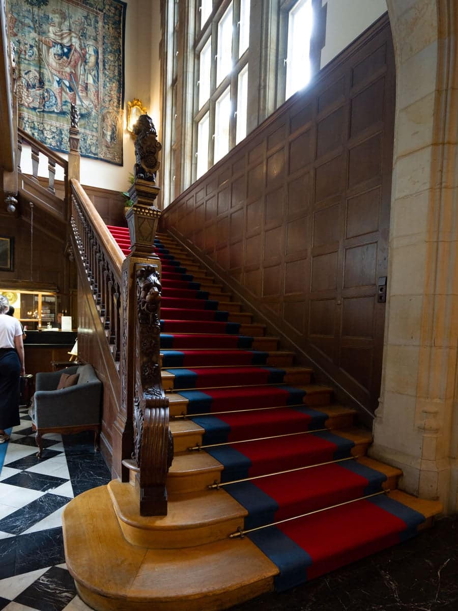 Eine prächtige Holztreppe mit rotem und blauem Teppichläufer, kunstvollem Geländer und großen Fenstern bildet den Rahmen für die Präsentation eines eleganten Brautkleids in einem Raum mit getäfelten Wänden und kariertem Marmorfußboden.