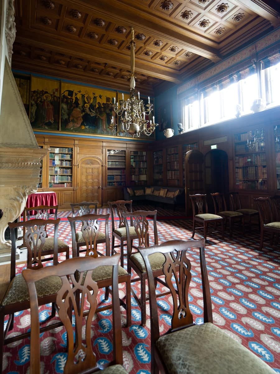 Ein reich dekorierter Bibliotheksraum mit verzierten Holzvertäfelungen, Bücherregalen, Kronleuchtern, gemusterten Teppichen und Reihen von leeren Holzstühlen vor einem Kamin und großen Fenstern - ein elegantes Ambiente, das sich perfekt für die Präsentation der Brautkleid-Stile eignet.