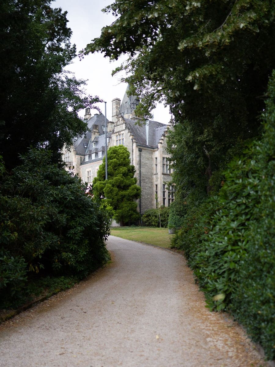 Ein von grünen Büschen und Bäumen gesäumter Kiesweg schlängelt sich zu einem großen Steingebäude mit hohen Dächern, das sich perfekt für schöne Brautkleid- oder Brautmoden-Fotos eignet, da es teilweise von üppigem Laub verdeckt wird.