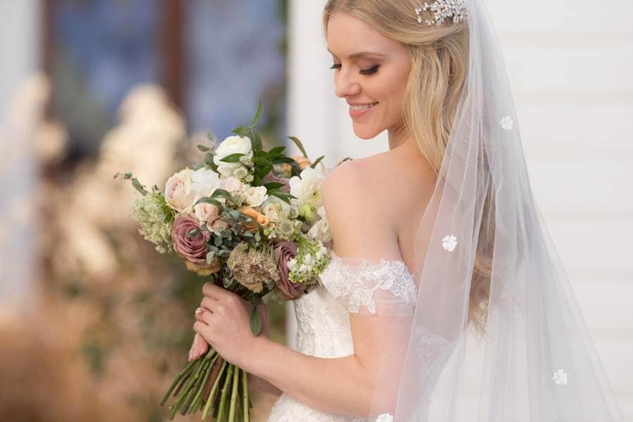 Eine lächelnde Braut in einem schulterfreien Spitzenhochzeitskleid mit langem Schleier hält einen Strauß pastellfarbener Blumen und blickt nach unten.